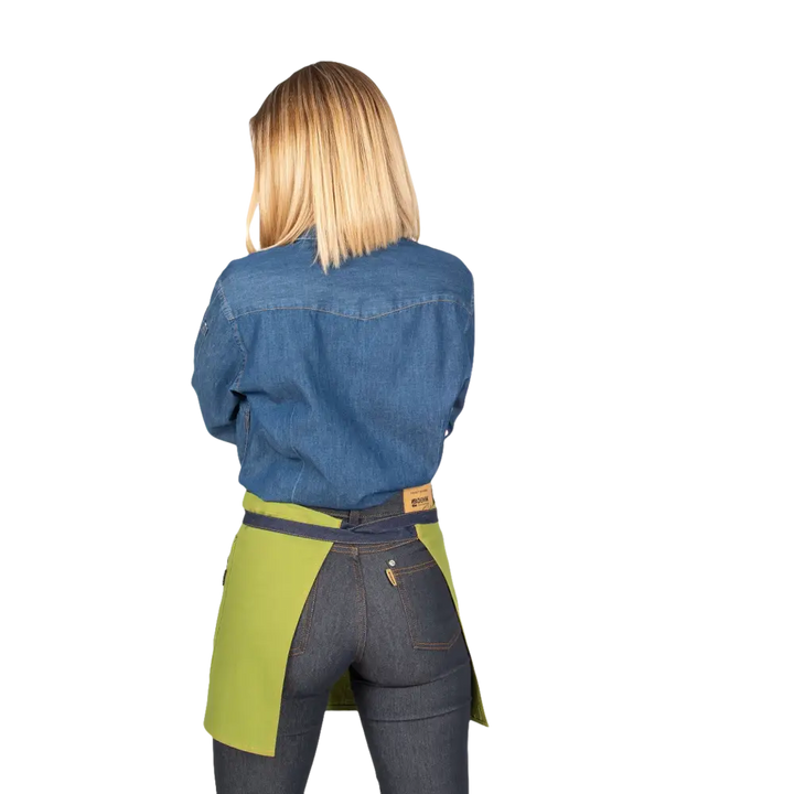 Donna bionda con camicia di jeans blu e grembiule bicolore verde e blu scuro su pantaloni scuri.